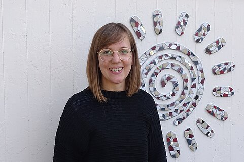 Porträt einer lächelnden Person mit schulterlangen braunen Haaren und runder Brille, in schwarzem Pullover, vor einer weißen Wand mit einer dekorativen Mosaiksonne.