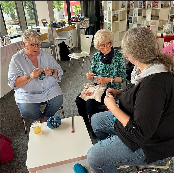 Drei Frauen sitzen in einem Gemeinschaftsraum und stricken gemeinsam, umgeben von Garn, Stricknadeln und einem kleinen Tisch mit Getränk.