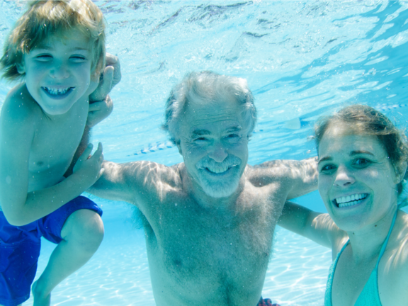 Unterwasseraufnahme: drei lächelnde Personen — ein Junge, eine Frau und ein älterer Mann — schwimmen gemeinsam in einem klaren Schwimmbad; der Mann hat die Arme um die anderen.