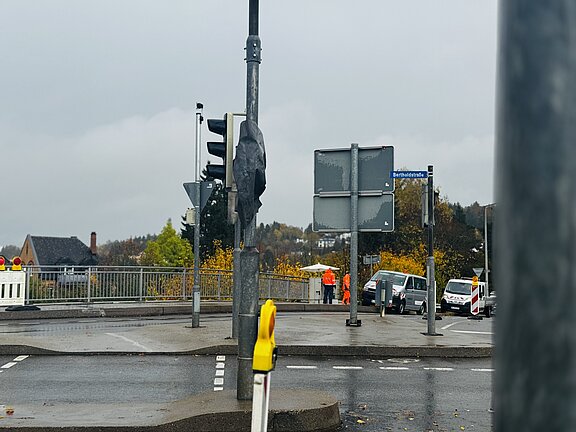 Nasse Straßenkreuzung an einer Brücke bei trübem Wetter: Ampel, Straßenschilder, Absperrungen, zwei Personen in orange Arbeitskleidung und mehrere Einsatzfahrzeuge am Fahrbahnrand.