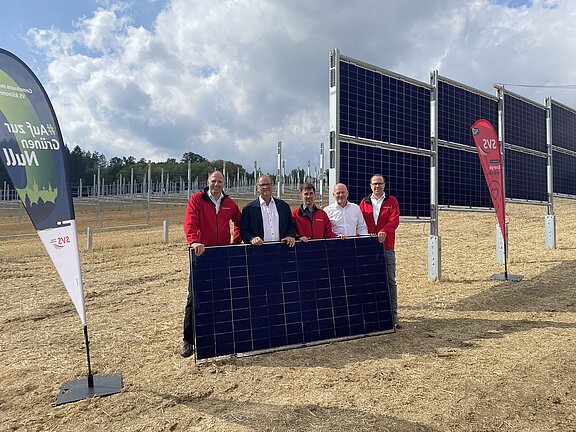 Fünf Personen stehen auf einem Feld vor mehreren aufgeständerten Solarmodulen und halten gemeinsam ein großes Solarpanel, offenbar zur Präsentation einer Photovoltaik-Anlage.