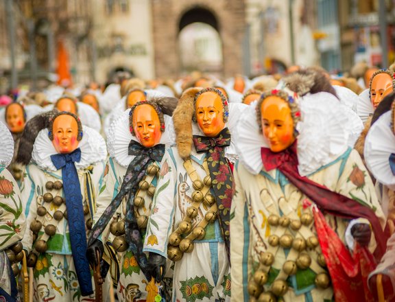 Viele verkleidete Menschen, welche vier Riemen mit Rollen um die Schuler tragen, laufen durch die Villinger Innenstadt. Jeder trägt eine Maske. 
