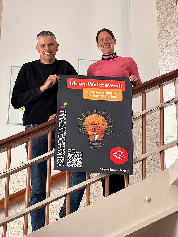 Zwei lächelnde Personen stehen auf einer Treppe und halten ein Plakat der Volkshochschule. Auf dem Plakat steht „Ideen‑Wettbewerb“ mit einer Glühbirnen‑Illustration und dem Aufruf, Kursideen einzureichen.
