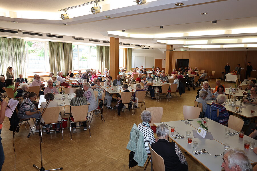 Heller Gemeindesaal mit vielen älteren Menschen an mehreren Tischen, die zusammen essen, trinken und sich unterhalten.