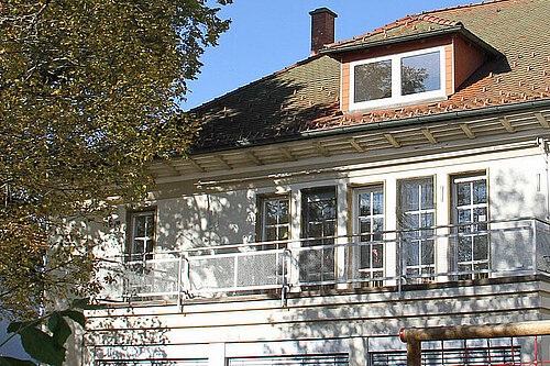 Weiße zweistöckige Villa mit Balustraden-Balkon, zahlreichen Sprossenfenstern und kleinem Dachgaubenfenster, umgeben von herbstlich gefärbten Bäumen.