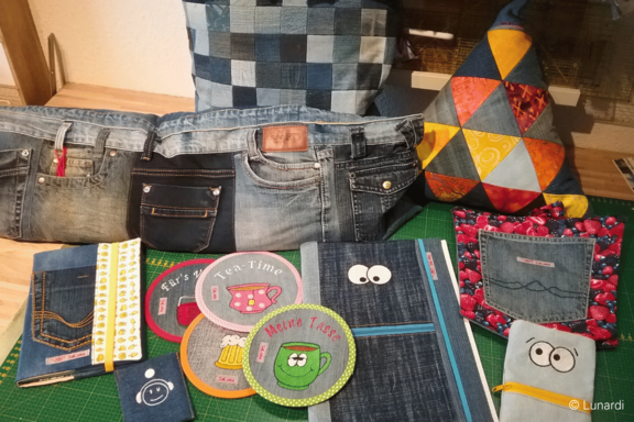 Verschiedene Upcycling-Objekte aus Jeansstoff auf einem Tisch: Taschen und Hüllen, genähte Untersetzer mit bunten Motiven, ein kleines Kissen mit aufgesetzter Jeans-Tasche und ein farbiges Patchwork-Kissen.