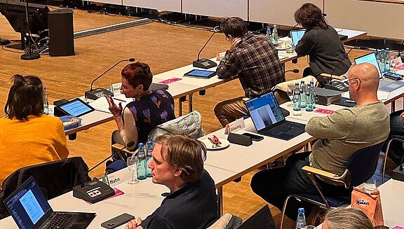 Großer Sitzungssaal mit vielen Teilnehmern an langen Tischen; Laptops, Mikrofone und Unterlagen sind sichtbar. Vorn eine Projektionsleinwand mit Präsentation — Szenenbild einer formellen Konferenz oder Versammlung.