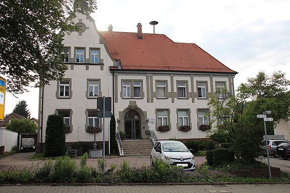 Historisches zweigeschossiges Verwaltungsgebäude mit rotem Ziegeldach, symmetrischer Fassade, Treppe zu einem Rundbogen-Eingang, Blumenkästen an den Fenstern und ein geparktes Auto im Vordergrund.