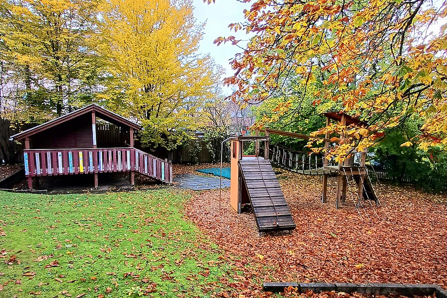 Kinderspielplatz im Herbst: rechts ein Klettergerüst mit Rutsche, links ein hölzernes Spielhaus/Veranda, umgeben von bunten Herbstbäumen und Laub auf dem Boden.
