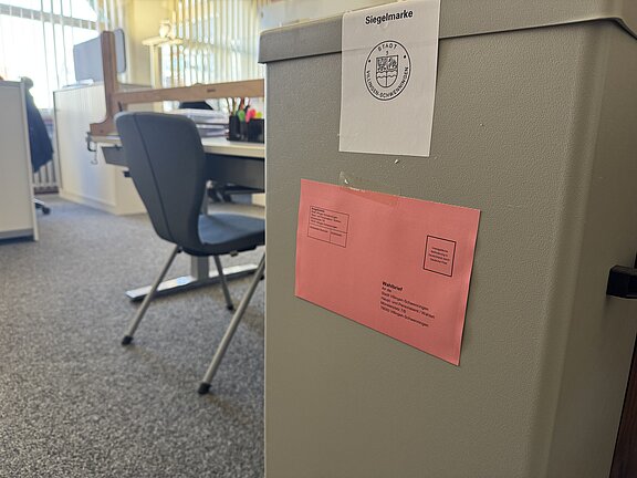Graue verschließbare Box (Wahlurne) in einem Büro, vorne ein rosa Umschlag angeklebt und ein Siegelaufkleber mit Behördenwappen darüber.