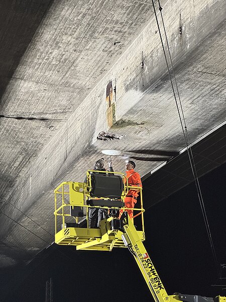 Zwei Bauarbeiter in orange Warnschutzkleidung auf einer gelben Hebebühne untersuchen einen beschädigten Bereich an der Unterseite einer Betonbrücke.