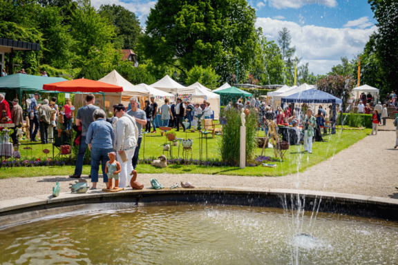Belebter Kunst- und Handwerksmarkt in einem Park: zahlreiche Besucher schlendern zwischen bunten Pavillons und Verkaufsständen, im Vordergrund ein Brunnen mit spritzendem Wasser.
