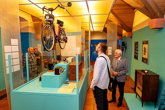 Drei Besucher in einem Museum betrachten ein an der Decke hängendes Fahrrad über einer Vitrine mit Retro-Objekten.