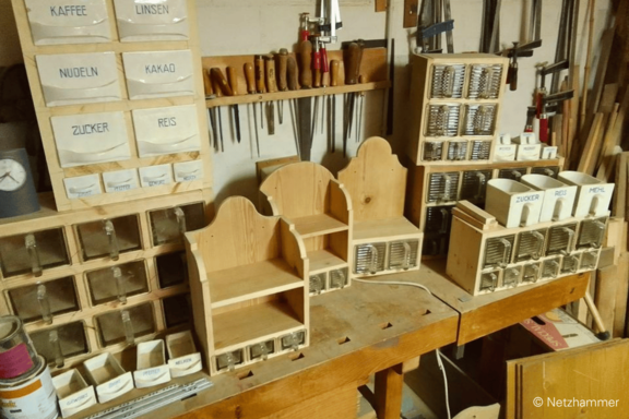 Holzwerkstatt mit handgefertigten Holzregalen und zahlreichen kleinen, beschrifteten Schubladen (Kaffee, Linsen, Nudeln, Kakao, Zucker, Reis) auf einer Werkbank; Werkzeuge hängen an der Wand.