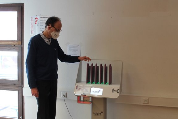 Ein Mann mit Mund-Nasen-Schutz steht vor einer Ladestation, in der mehrere tabletähnliche Geräte senkrecht in Fächern mit grünen Leuchten aufgeladen werden.