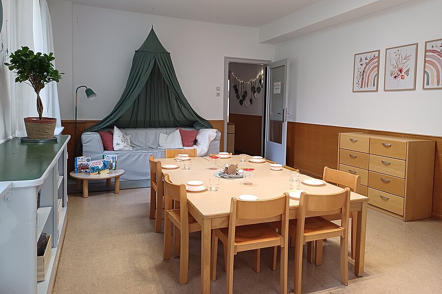 Heller Gruppenraum mit großem rechteckigem Holztisch und neun kleinen Holzstühlen, gedeckt mit Tellern. Dahinter Sofa mit grünem Baldachin, Kommode und Wandbildern.