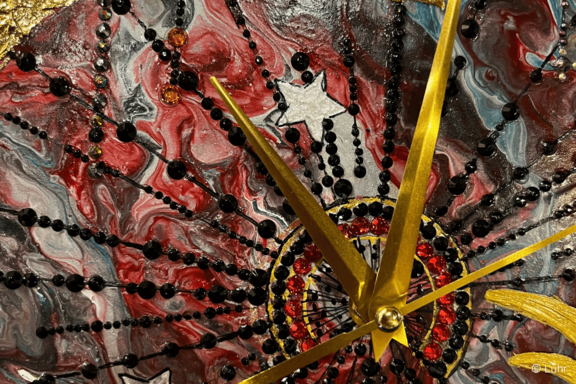 Nahaufnahme einer dekorativen Uhr: goldene Zeiger auf einem roten, schwarzen und grauen marmorierten Zifferblatt mit strahlenförmigen Perlen- und Strassverzierungen sowie kleinen Sternmotiven.