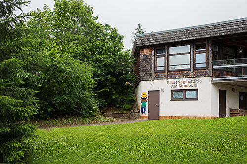 Eingang einer Kindertagesstätte "Am Kopsbühl" in einem zweistöckigen Gebäude neben Bäumen, mit bemaltem Kinderfigurenmotiv an der Tür und einer Rasenfläche im Vordergrund.