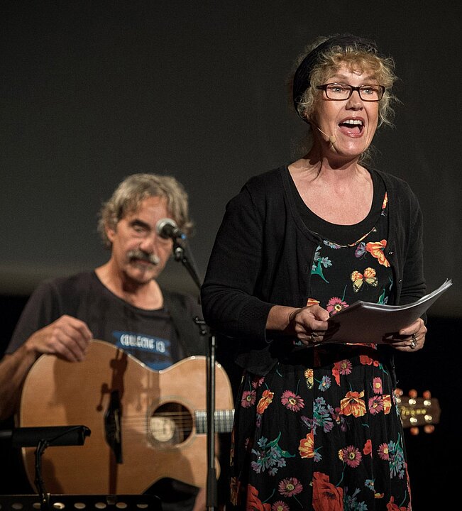 Zwei Personen auf einer Bühne: links ein Mann mit Gitarre, rechts eine Frau mit Mikrofon und Blumenkleid. (KI generiert)