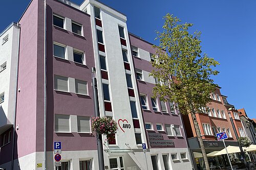 Rosa-weißes fünfstöckiges Gebäude der Arbeiterwohlfahrt (AWO) an sonniger Innenstadtstraße, mit AWO-Herzlogo an der Fassade, Eingangsbereich, Parkplätzen und einem Baum davor.