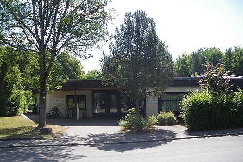 Flaches, eingeschossiges Gebäude mit überdachtem Haupteingang, Fahrrädern neben der Tür, umgeben von Bäumen und Sträuchern; Straße im Vordergrund.
