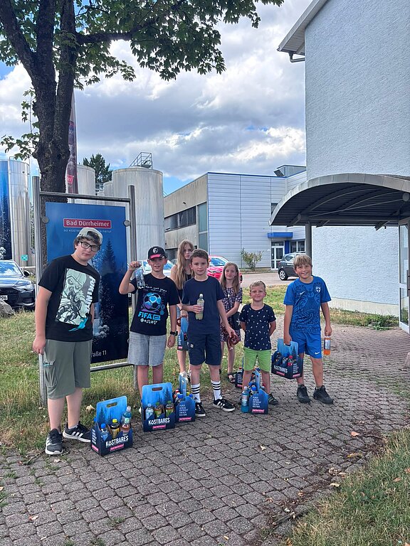 Sieben Kinder und Jugendliche stehen draußen vor einem Gebäude, lächeln in die Kamera und halten beziehungsweise stellen jeweils Getränkekästen mit Flaschen ab.
