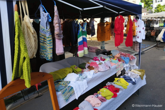 Marktstand mit handgestrickten und gehäkelten Artikeln: bunte Schals, Mützen, Pullover, Babybekleidung und Decken auf Tischen und an Kleiderstangen.