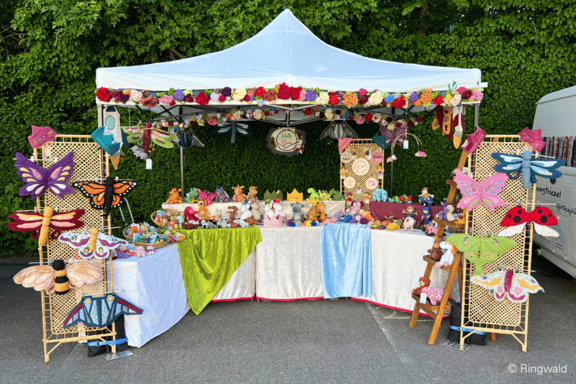Marktstand unter weißem Pavillon, geschmückt mit Blumen, zeigt bunte handgefertigte Stofftiere, Holzschmetterlinge, Marienkäfer und andere verspielte Deko auf Tischen.