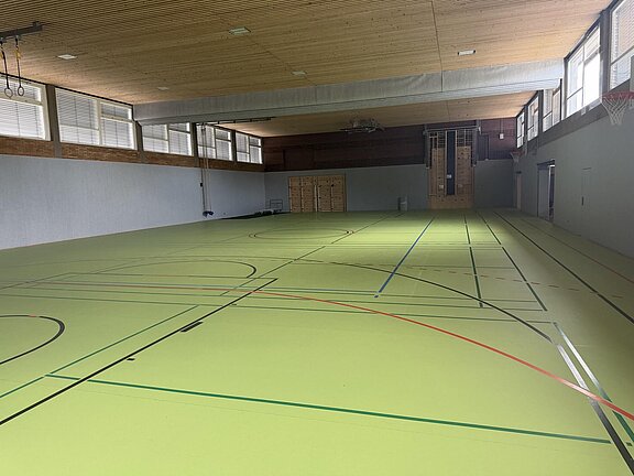 Leere Sporthalle mit hellgrünem Hallenboden, farbigen Spielfeldlinien, hoher Holzdecke, seitlichen Fenstern und einem Basketballkorb an der rechten Wand.