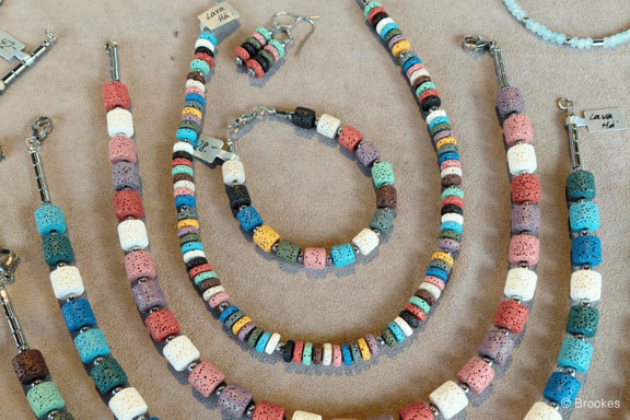 Mehrere bunte Halsketten, ein Armband und Ohrringe aus würfelförmigen, porösen Lavastein-Perlen in Pastell- und kräftigen Farbtönen, auf beigem Stoff ausgebreitet.