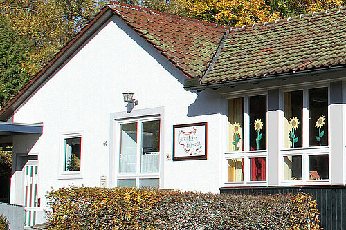 Eingeschossiges weißes Gebäude mit langem Fensterband, hinter dem bunte Kinderzeichnungen hängen; kleiner Vorplatz und herbstliche Bäume im Hintergrund.
