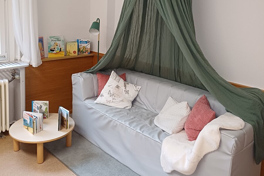 Gemütliche Leseecke mit hellgrauem Sofa unter grünem Baldachin, mehreren Kissen und Decke, kleinem runden Tisch mit aufgestellten Kinderbüchern und einer Leselampe.