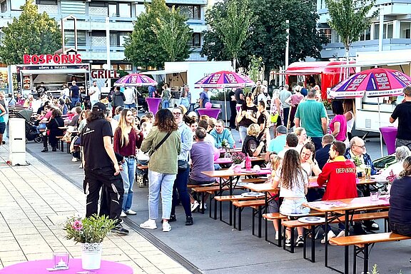 Belebtes Open-Air-Food-Festival: viele Menschen sitzen an langen Holzbänken mit bunten Sonnenschirmen, umgeben von Imbissständen und Foodtrucks.