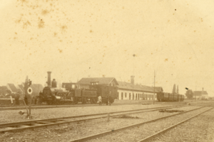 Sepiafoto eines historischen Bahnhofs mit mehreren Gleisen, einer Dampflokomotive, einem Empfangsgebäude und wartenden Bahnwagen.
