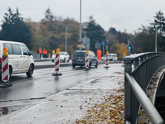 bertholdbrücke Nasse Straße mit Baustellenabsperrungen und Warnleuchten, Fahrzeuge passieren eine einspurige Baustelle; Bauarbeiter in orange Jacken im Hintergrund; Laub am Fahrbahnrand neben einem Brückengeländer.