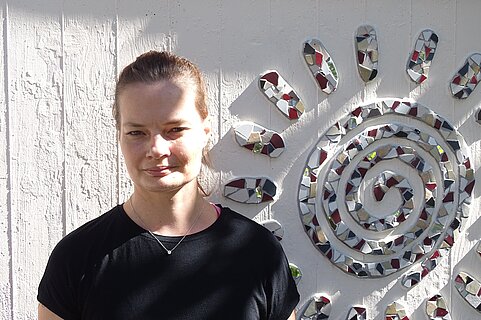 Junge Frau in schwarzem T‑Shirt steht vor einer weißen Wand; rechts daneben eine große Spiral‑Mosaiksonne aus bunten, spiegelnden Stücken.