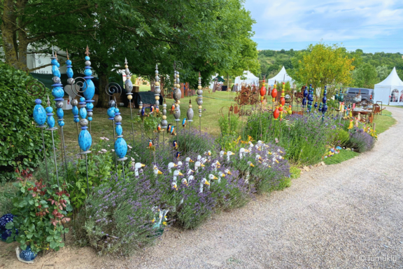 Reihe bunter Keramik-Gartenstecker in Kugel- und Tropfenformen zwischen Lavendel und Blumen an einem Kiesweg, im Hintergrund Wiese, Bäume und weiße Pavillons.