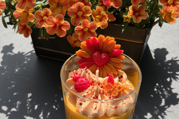 Glas mit gelber Cremeschicht, darauf weiße Sahnetupfen und rote‑orange essbare Dekorationen: blumenförmige Garnitur, kleines Herz, rote Praline und Streusel.