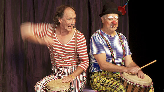 Zwei fröhliche Clowns auf einer Bühne spielen Handtrommeln; links rot-weiß gestreiftes Shirt, rechts blau-weiß gestreiftes Shirt, gelb-kariere Hose, rote Clownsnase und Hut.