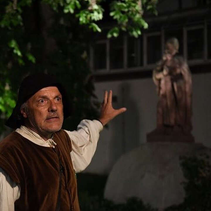 Ein Mann in mittelalterlicher Kleidung zeigt auf eine Statue im Hintergrund. (KI generiert)