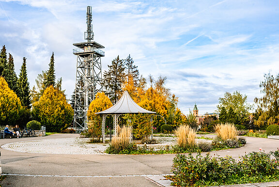 Park im Herbst mit metallischem Aussichtsturm, kleinem Pavillon und geschwungenen Wegen, umgeben von bunten Laubbäumen und Ziergräsern.