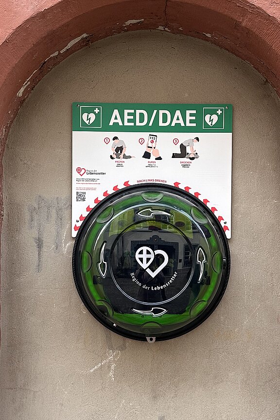 In einer bogenförmigen Nische an einer Hauswand hängt ein rundes grünes Gehäuse mit einem automatischen externen Defibrillator (AED), beschildert "AED/DAE".