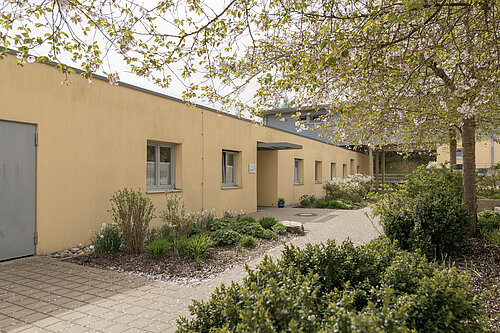 Einfache eingeschossige gelbe Gebäudezeile mit Eingangsüberdachung, Gehweg und gepflegten Blumenbeeten sowie Büschen unter Baumkronen in einem ruhigen Innenhof.