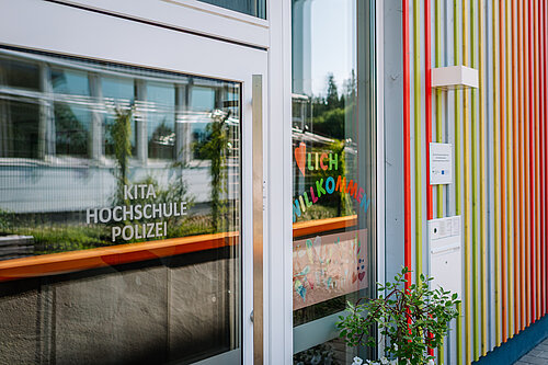 Die Eingangstür einer Einrichtung mit den Aufschriften „KITA“, „HOCHSCHULE“ und „POLIZEI“. Bunte Buchstaben auf dem Fenster begrüßen mit „HERZLICH WILLKOMMEN“. Im Vordergrund eine kleine Pflanze. Das Gebäude hat eine farbige Holzfassade.