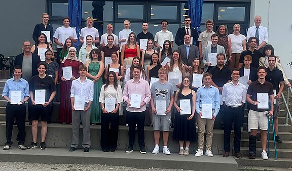 Gruppenfoto von vielen Personen auf Treppenstufen; die meisten halten Zertifikate und lächeln. Unterschiedliche Kleidung von festlich bis leger — Abschluss- oder Ehrungsfeier.