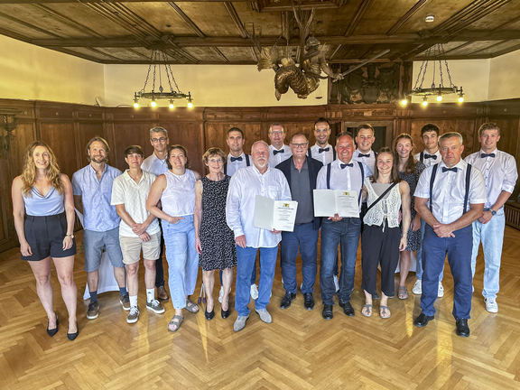 Gruppenfoto von rund 18 Personen in einem Holzsaal; in der Mitte halten zwei Männer Urkunden, die Gruppe lächelt.