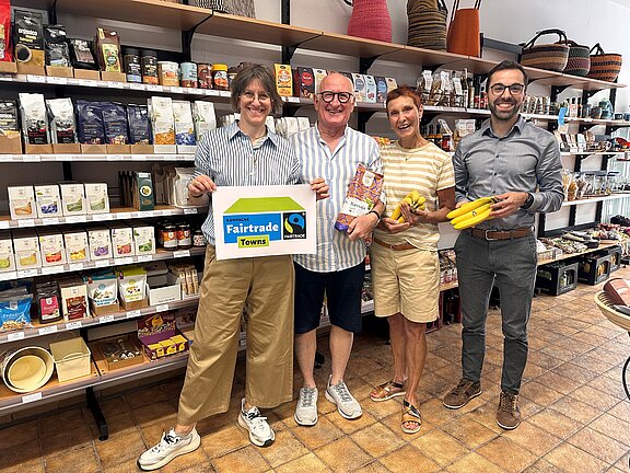Vier lächelnde Personen in einem Laden halten ein Fairtrade-Schild sowie Bananen und Fairtrade-Produkte vor Regalen mit Lebensmitteln.