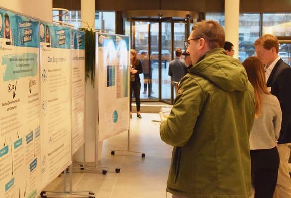 Mehrere Menschen in einem Eingangsbereich lesen Informationsplakate auf Stelltafeln bei einer Ausstellung oder Veranstaltung.