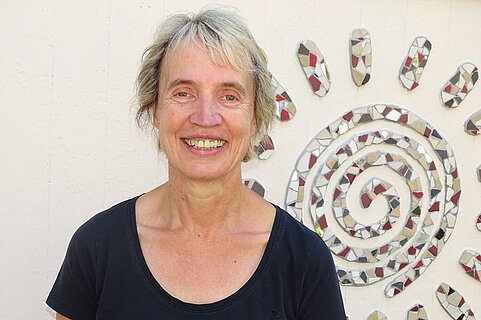 Frau mittleren Alters mit kurzem blondem Haar und breitem Lächeln, trägt schwarzes T‑Shirt, vor einer Wand mit dekorativem Mosaik‑Sonnenmotiv.