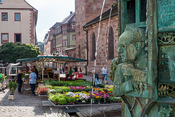 Marktplatz mit bunten Blumenständen und flanierenden Menschen; im Vordergrund bronzene Wasserfigur eines nachdenklichen Mannes, aus deren Mund Wasser läuft.